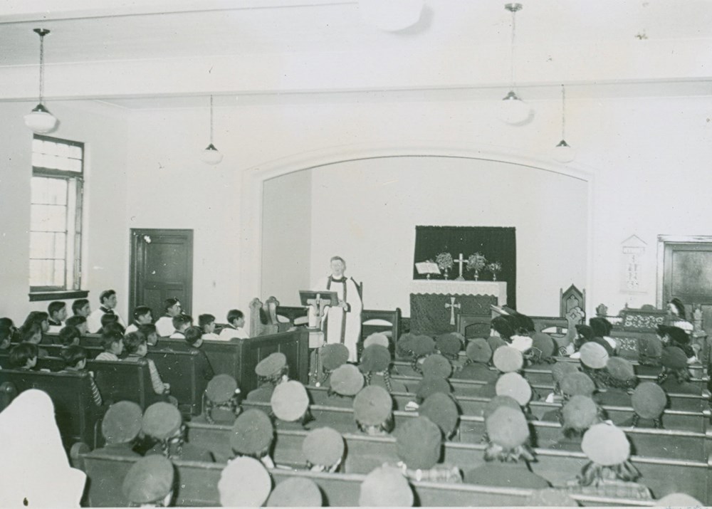 Morning Prayer. View from rear to front of school chapel, with pews filled with students and distant principal conducting service- [194-?]. P75-103-S7-185 from The General Synod Archives, Anglican Church of Canada