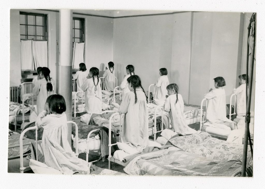 Jr. Girls' Dormitory during the filming of a documentary, with the addition of window coverings and nicer quilts on the beds. Old Sun IRS, 1952. Photo Source: Shingwauk Residential Schools Centre, Algoma University