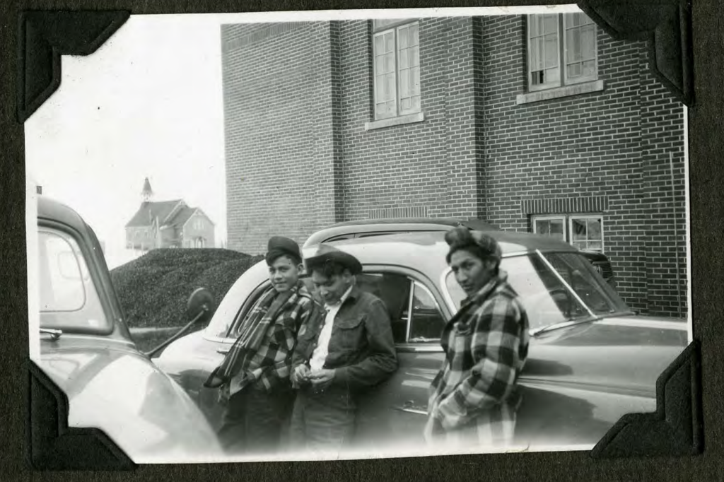 Boys in parking lot outside of the boys' playroom exit. Chapel behind them, with former church in the background. 1950s. Shingwauk Residential Schools Centre, Algoma University