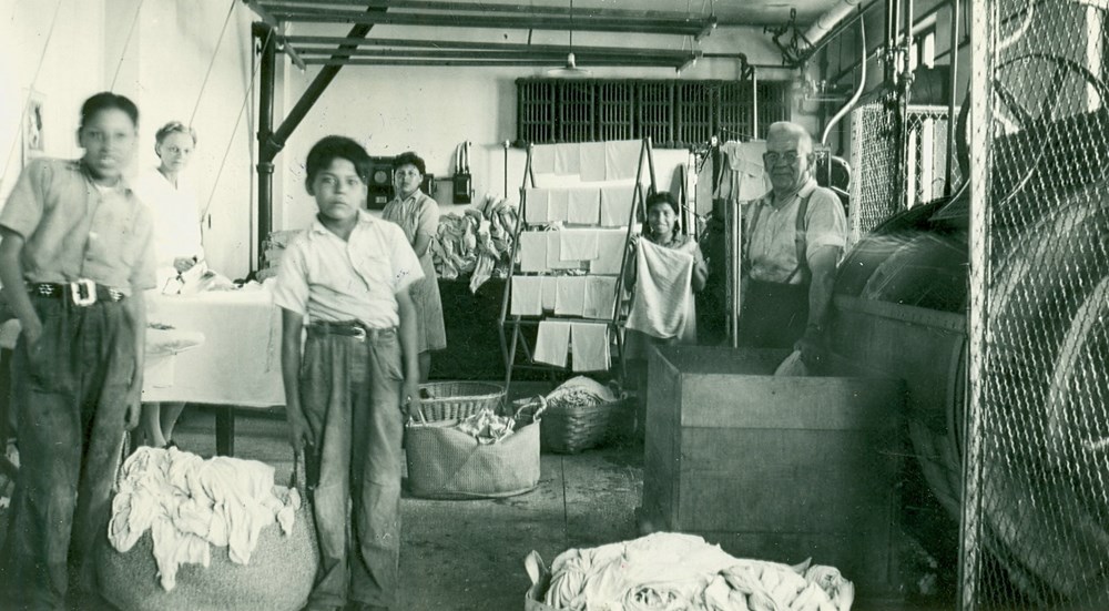 Laundry work in progress, involving students Hilda Lowhorn, Edna Studhorse, Ronald Cutter, Wallace Bearchief and staff members Mrs. J. Champness (Laundry Matron) and Mr. W.H. Palmer (Engineer?). 1945. P75-103-S7-168 from The General Synod Archives, Anglican Church of Canada