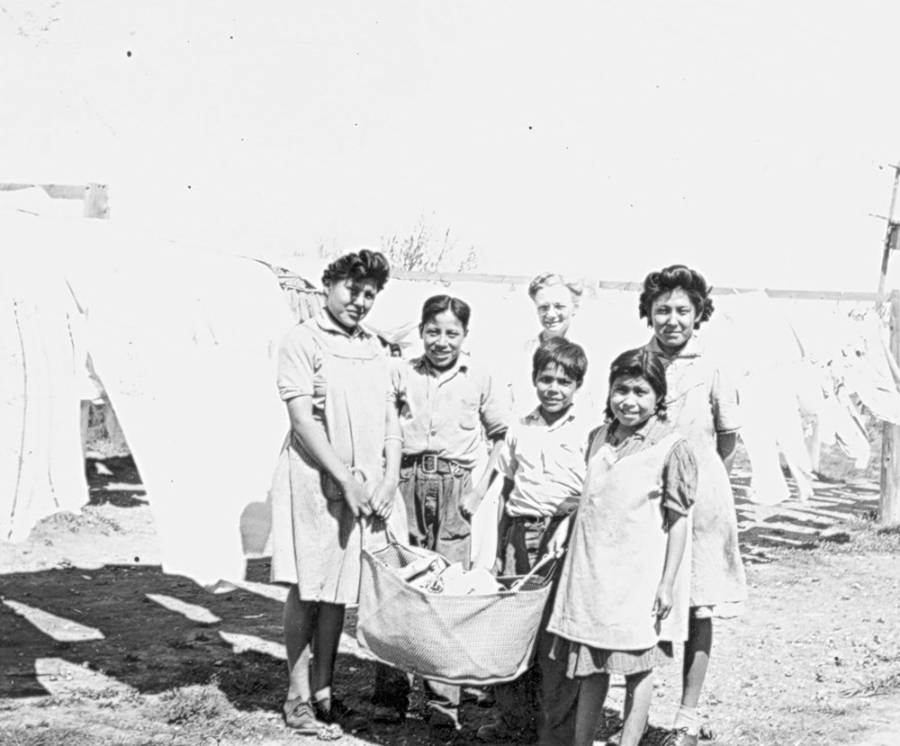 Students Hilda Lowhorn, Ruby Simson, Edna Studhorse, Ronald Cutter and Wallace Bearchief assist Laundry Matron Mrs. J. Champness to hang clothing on the line, outside school. 1945. P75-103-S7-170 from The General Synod Archives, Anglican Church of Canada