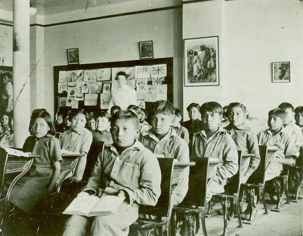 Junior Classroom. Posed photo of students seated at their desks with teacher (blurred) standing at the back - 1945. P75-103-S7-182 from The General Synod Archives, Anglican Church of Canada