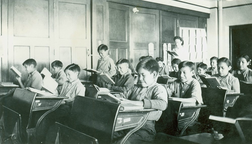 Grades 3 and 4. View of classroom with students at their desks and teacher standing at rear- [194-?]. P75-103-S7-193 from The General Synod Archives, Anglican Church of Canada