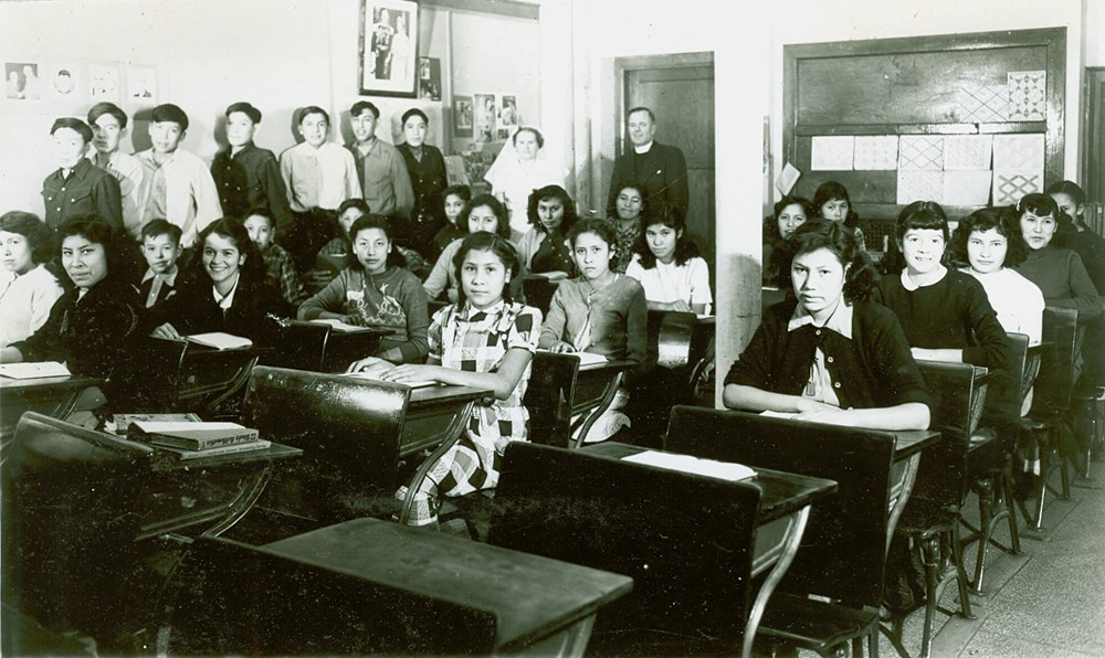 Senior students. Photo consists of senior girls at their desks, boys standing against side wall, and Principal and staff member standing at rear- [194-?]. P75-103-S7-201 from The General Synod Archives, Anglican Church of Canada