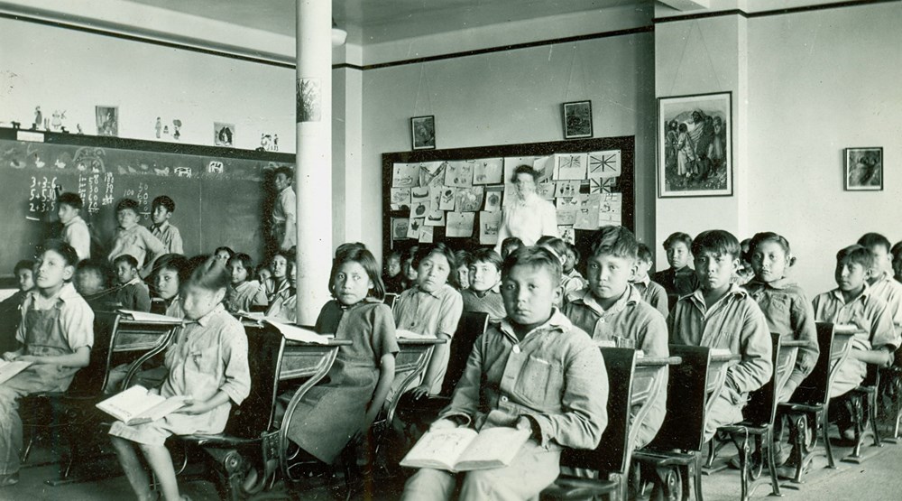 Junior Classroom. Posed photo of students seated at their desks with teacher (blurred) standing at the back - 1945. P7538-1005 from The General Synod Archives, Anglican Church of Canada