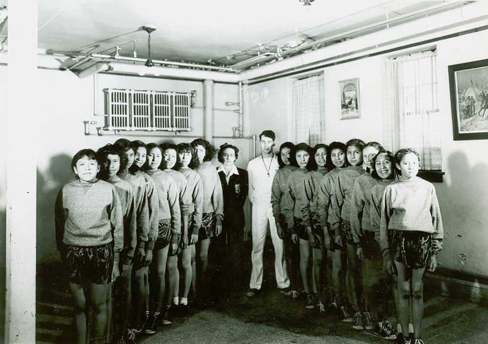 Old Sun School, Gleichen, Alberta - Girls' sports team and coaches, taken in student dining room - [194-?]. P7538-1013 from The General Synod Archives, Anglican Church of Canada