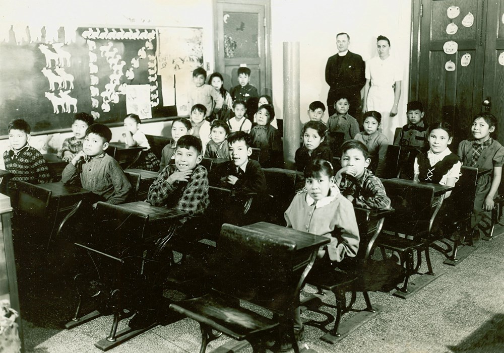 Formal classroom photo of students at their desks. Principal and teacher stand at the back - [194-?]. P7538-1015 from The General Synod Archives, Anglican Church of Canada