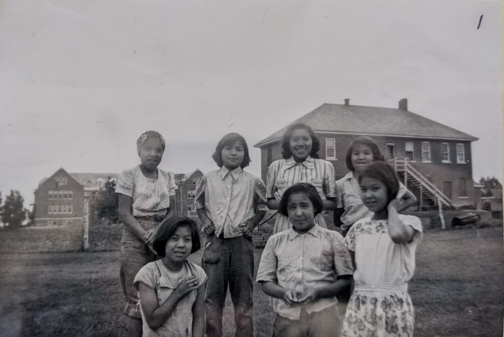 Grade 4 and 5 students standing at the back of the carriage house with the main school building visible behind them, 1926-1937. PR1985.0100 from The Provincial Archives of Alberta, Open Copyright.