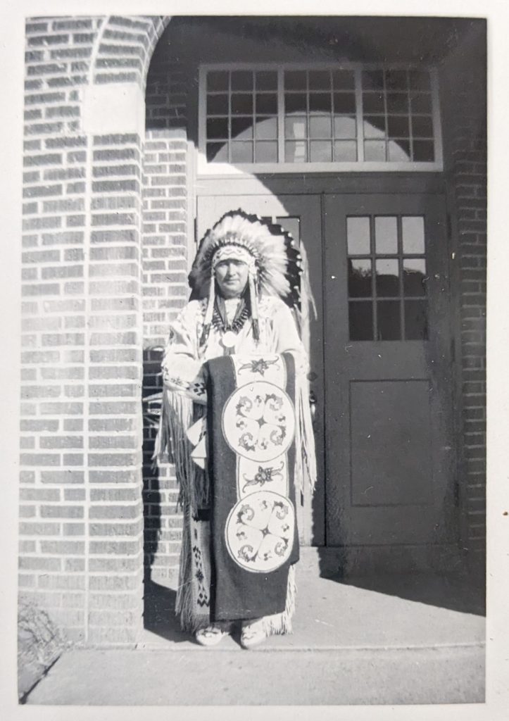 Man dresses in regalia on front steps of Blue Quills, 1947-1951. PR1973.0248/864 from The Provincial Archives of Alberta, Open Copyright.