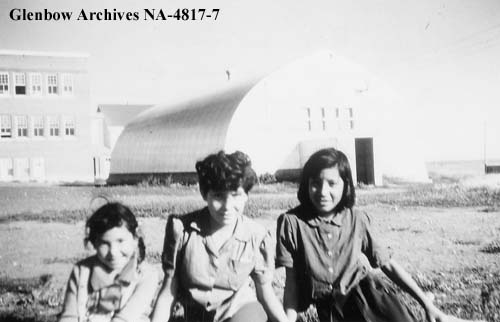 Sylvia Peacemaker sits between two friends in front of a quonset hut used for physical education. 1955-1957. NA-4817-7. Courtesy of Glenbow Archives.